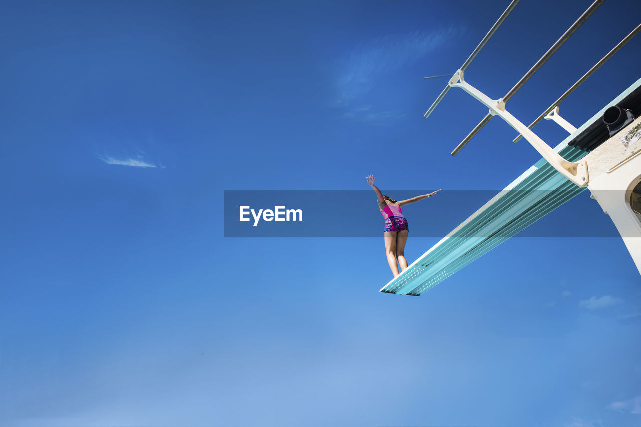 Low angle view of carefree girl standing on diving platform against blue sky during sunny day
