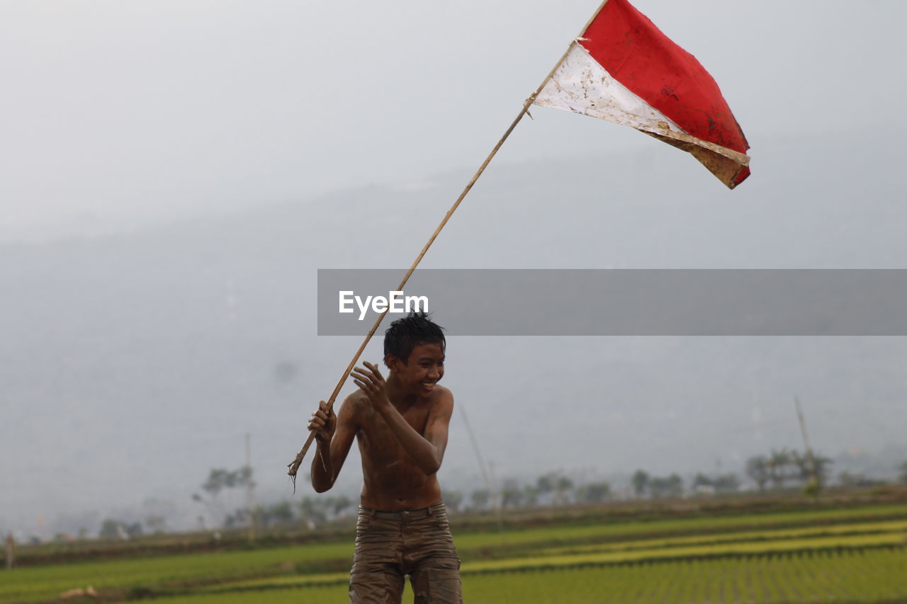 Shirtless boy holding flag while standing at farm against cloudy sky