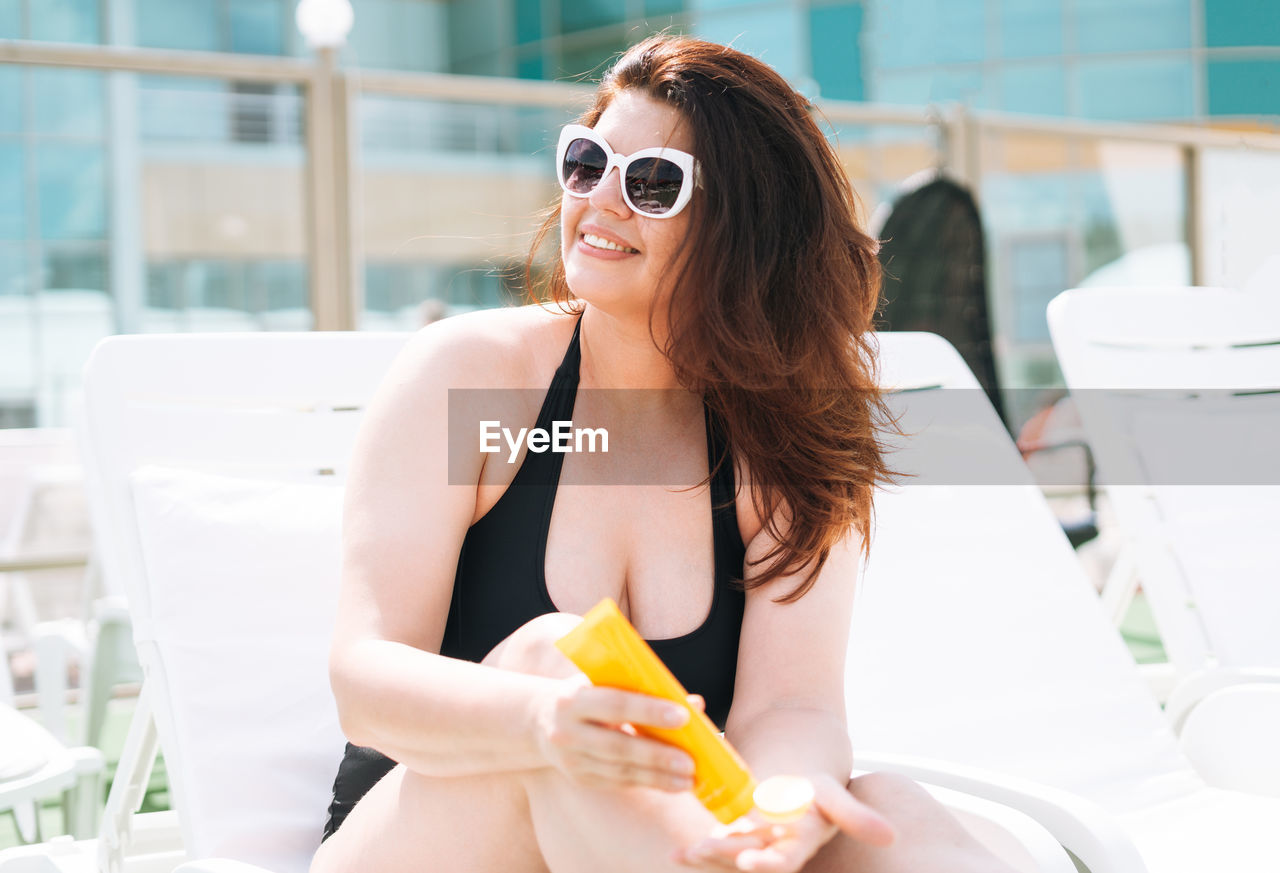 Stylish woman plus size in black swimsuit with sunscreen cream in hands on beach lounger near pool