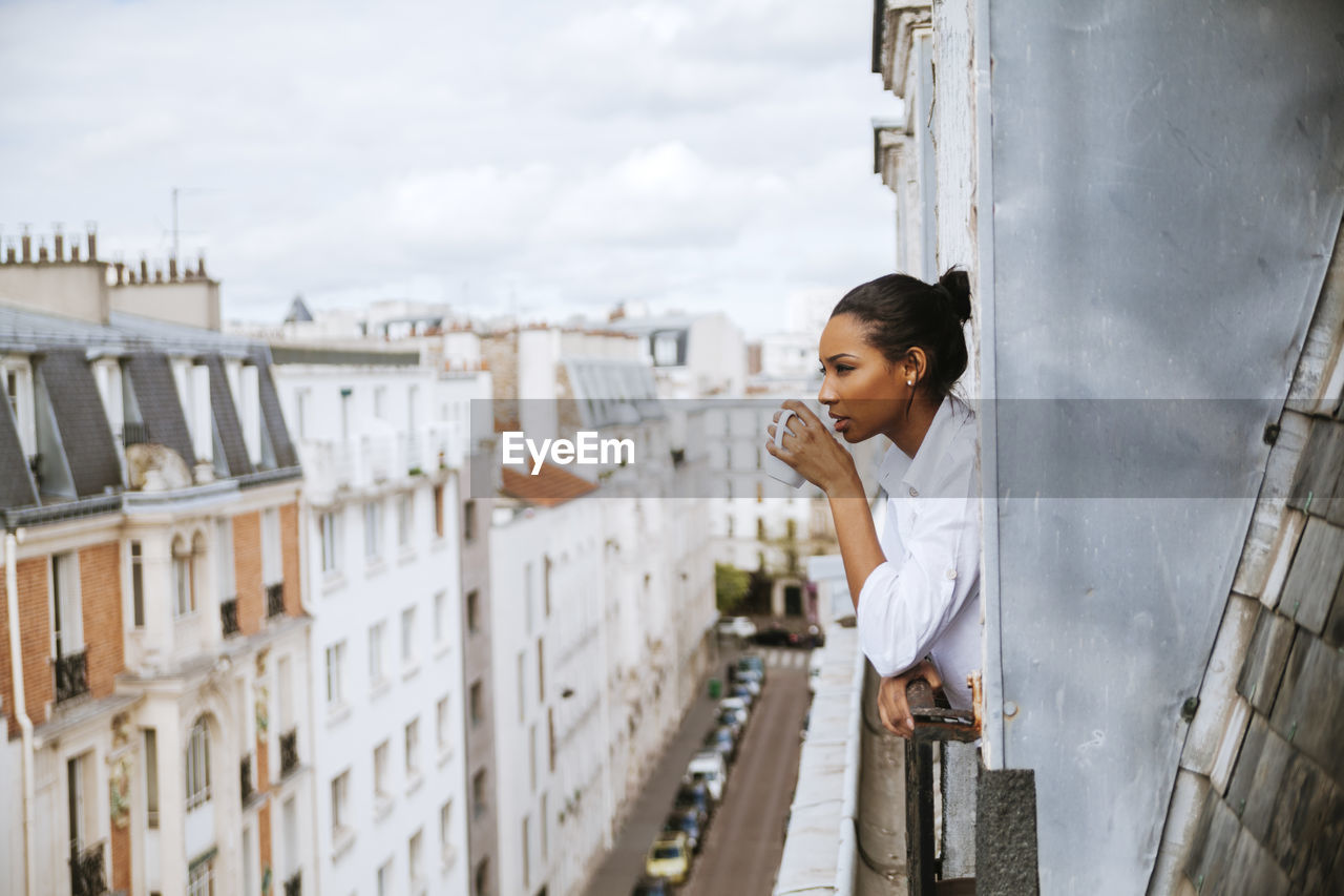 France, paris, young woman with cup of coffee leaning out of window