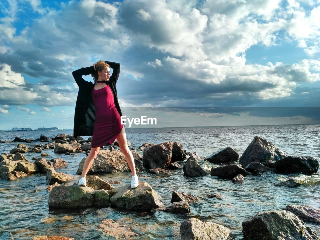 Young woman posing on rocks in sea against cloudy sky