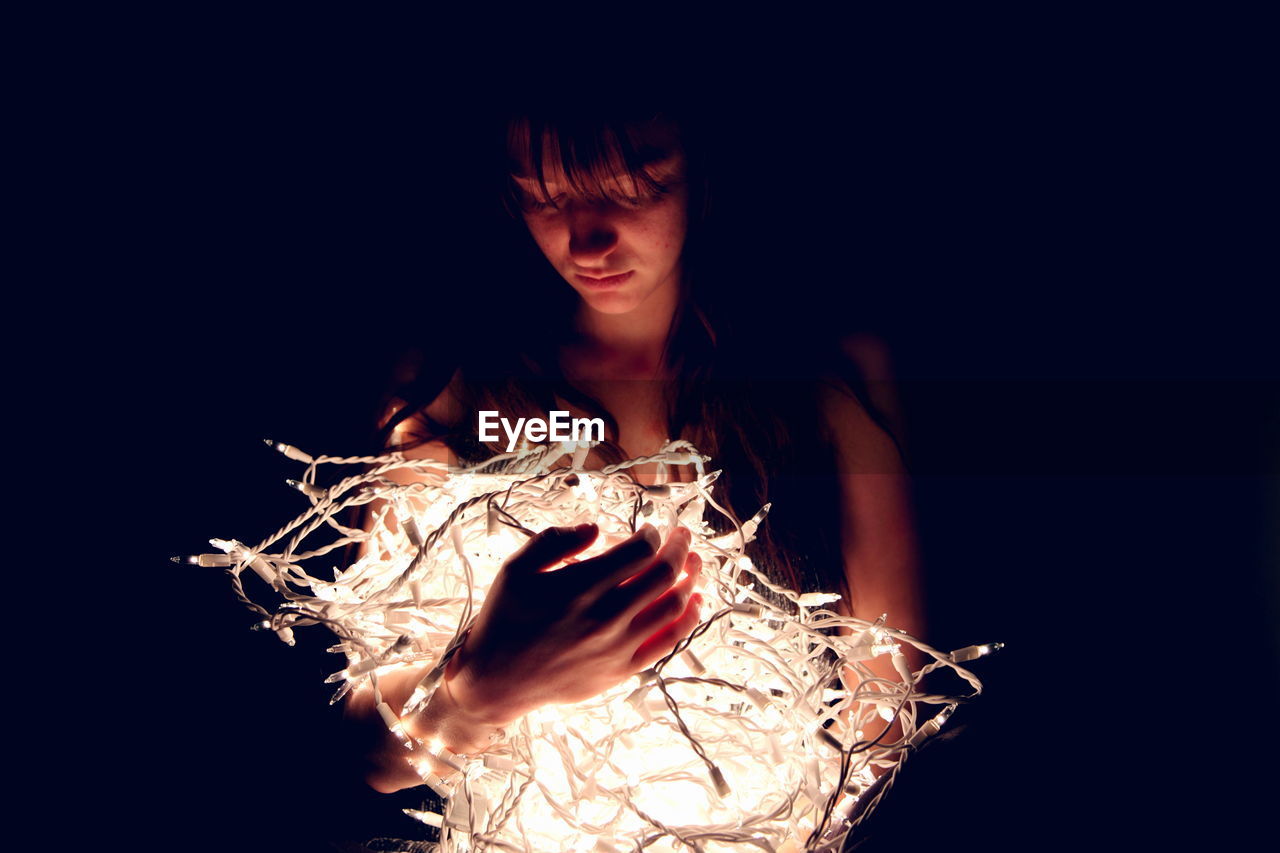 Close-up of teenage girl with illuminated string lights against black background