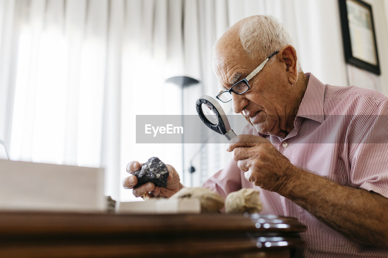 Retired senior male using magnifying glass for research of fossil and mineral at home