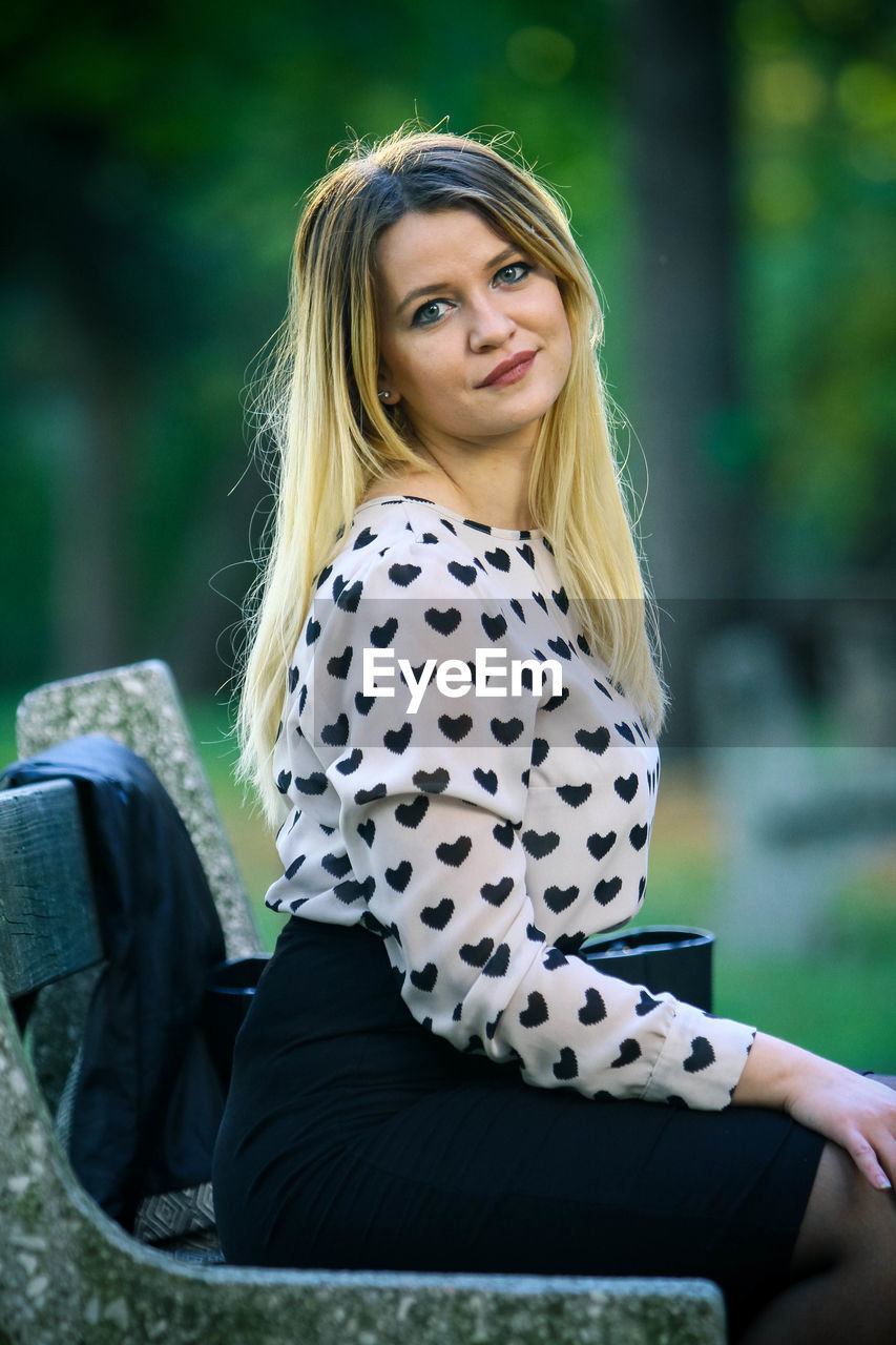 Portrait of smiling young woman sitting on bench at public park