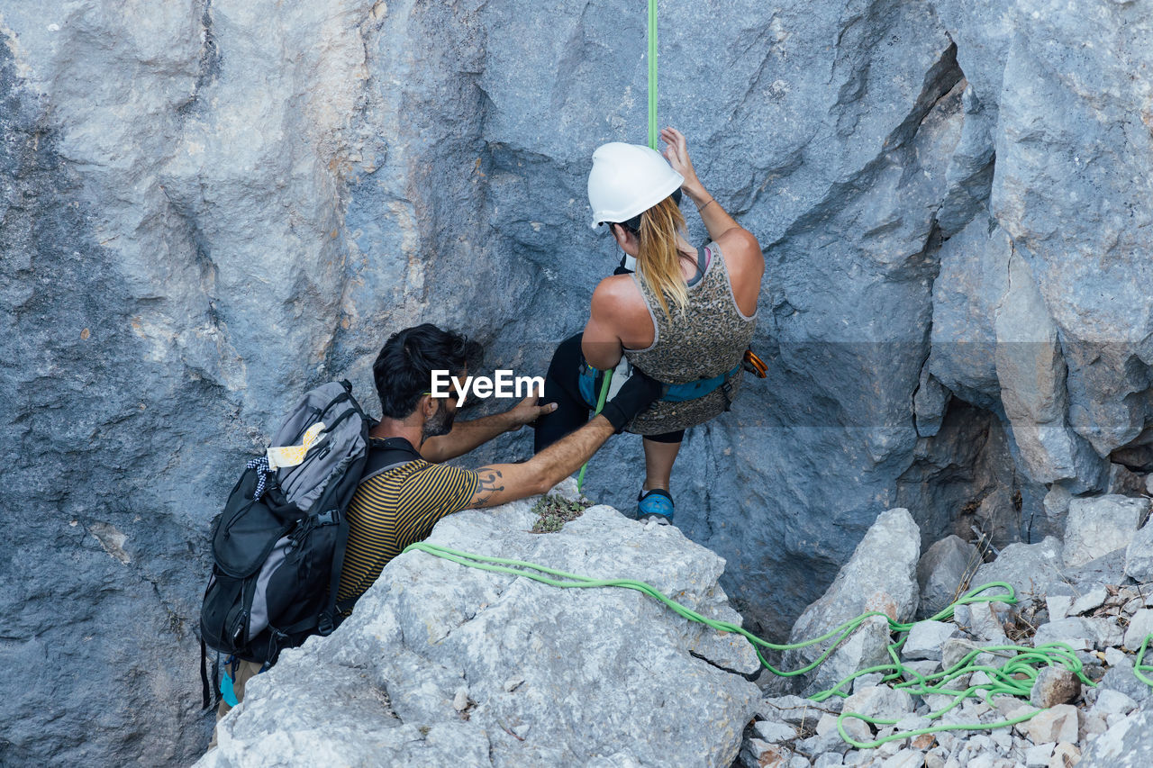 Confident female in safety harness and helmet getting ready for ascending and listening to instructions of male friend while practicing climbing together in mountains