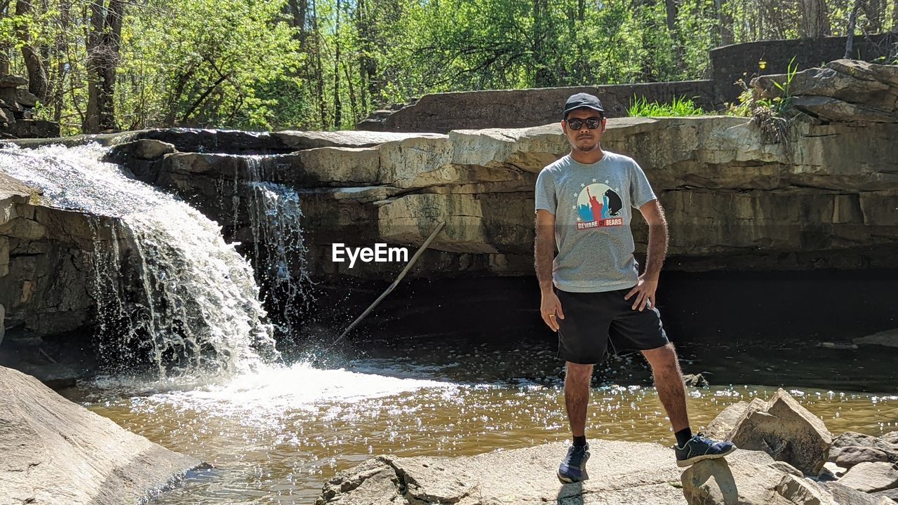 one person, water, full length, nature, lifestyles, adult, men, front view, waterfall, standing, leisure activity, day, casual clothing, tree, plant, young adult, motion, sports, shorts, outdoors, sunlight, rock, clothing, person, portrait, footwear, sunglasses, glasses, land, looking at camera