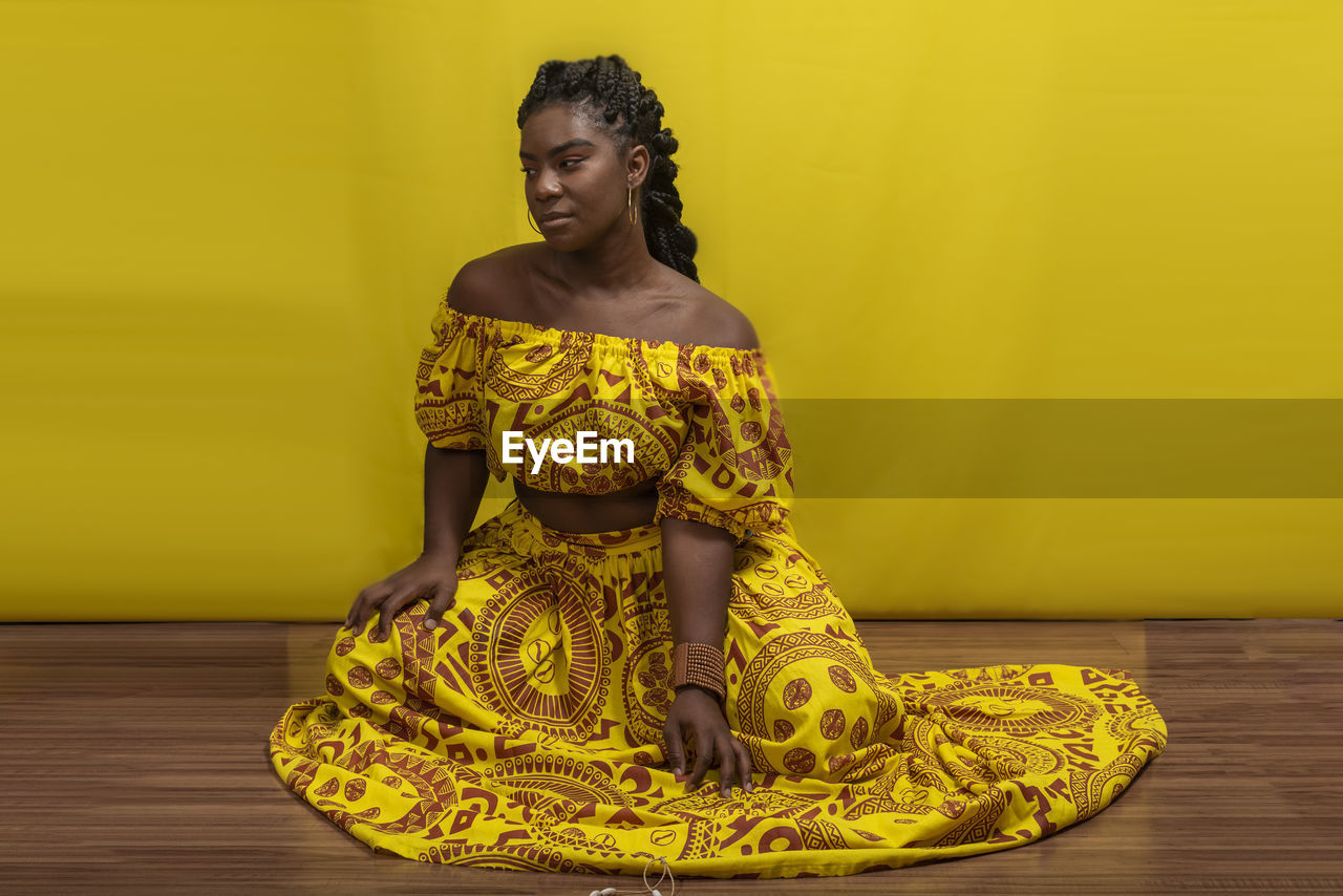 Young beautiful woman sitting on wooden floor, wearing colorful dress. against yellow background.