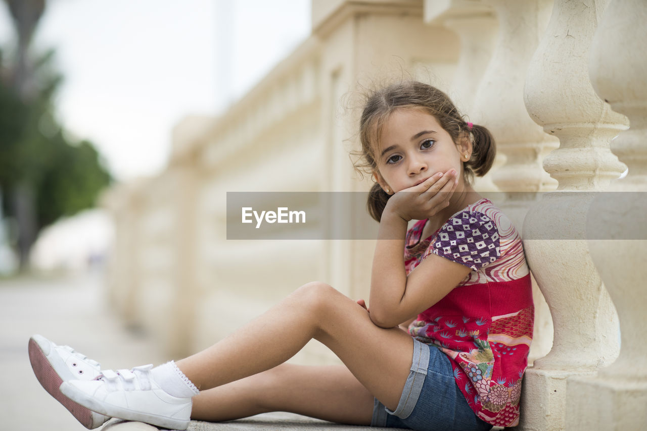 Portrait of cute girl sitting by railing in city