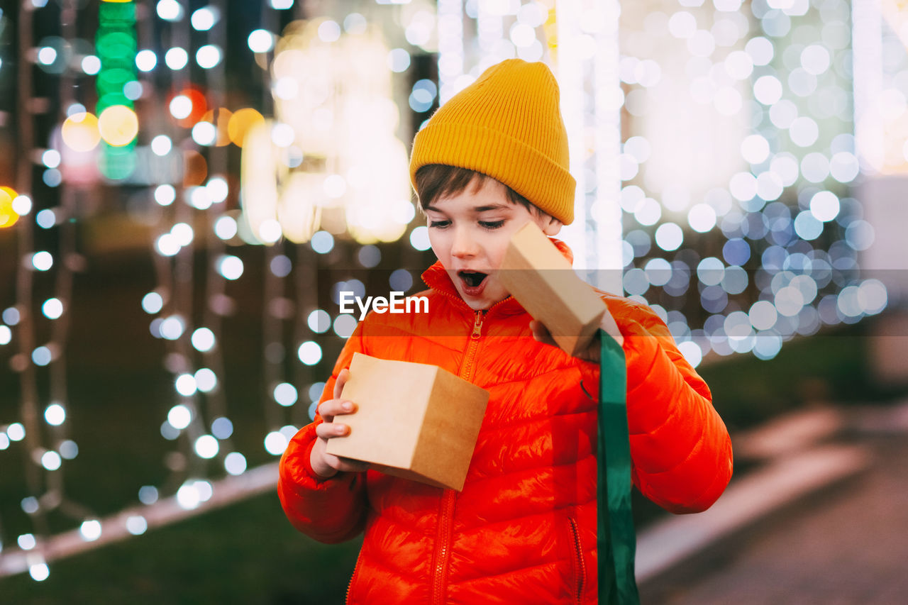 Portrait of a happy boy standing on the evening street and opening a christmas gift. 