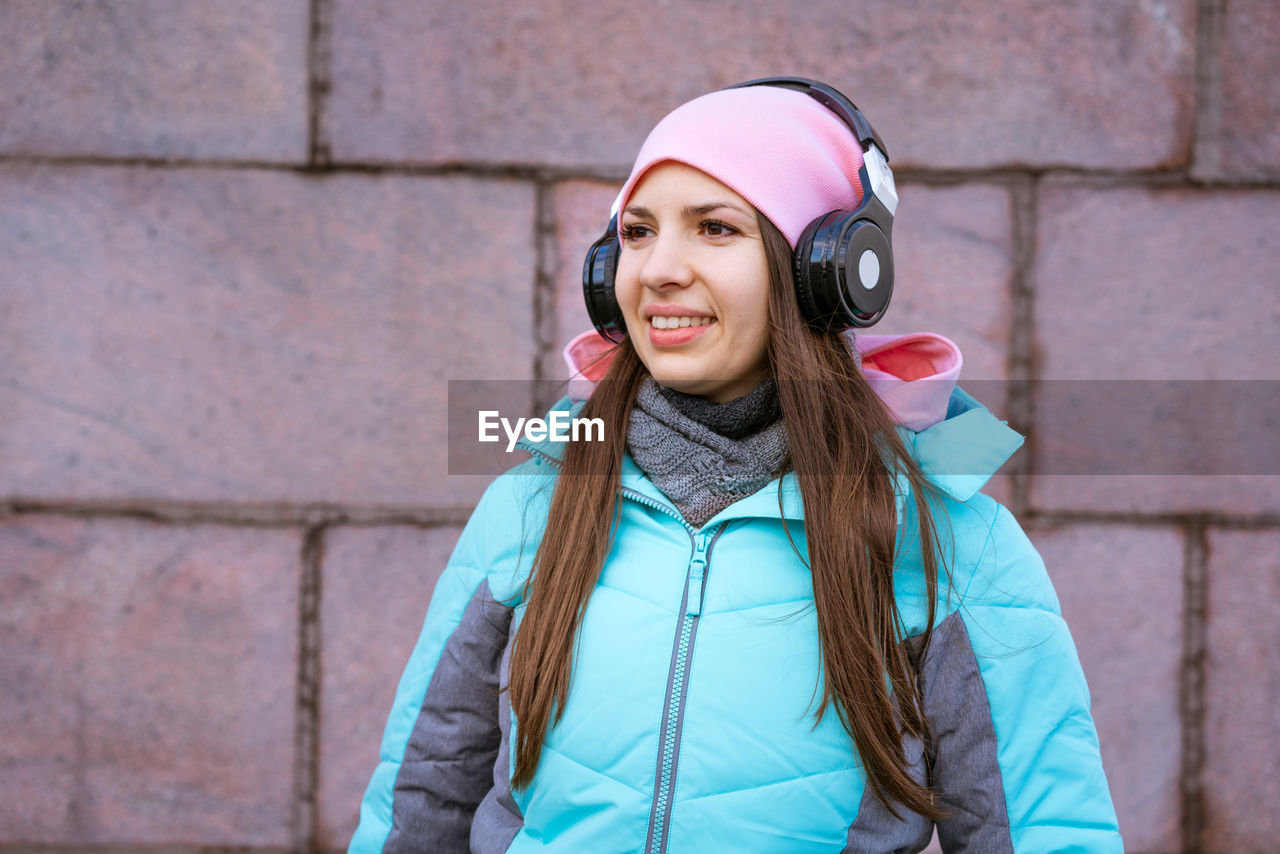 Happy young woman in warm jacket and pink hat listening to music on headphones