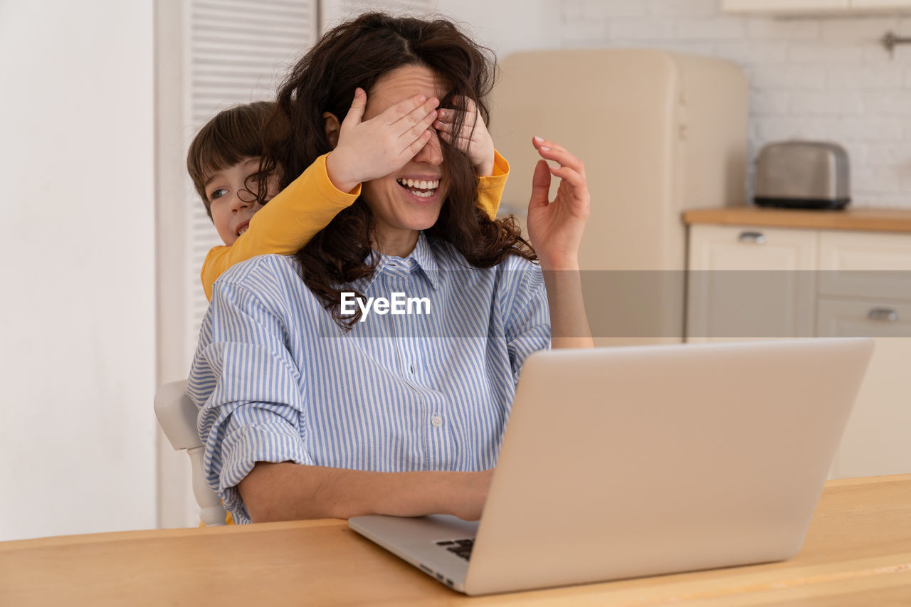 Mom work on laptop at home during lockdown, child distracts from work, covering her mother's eyes.