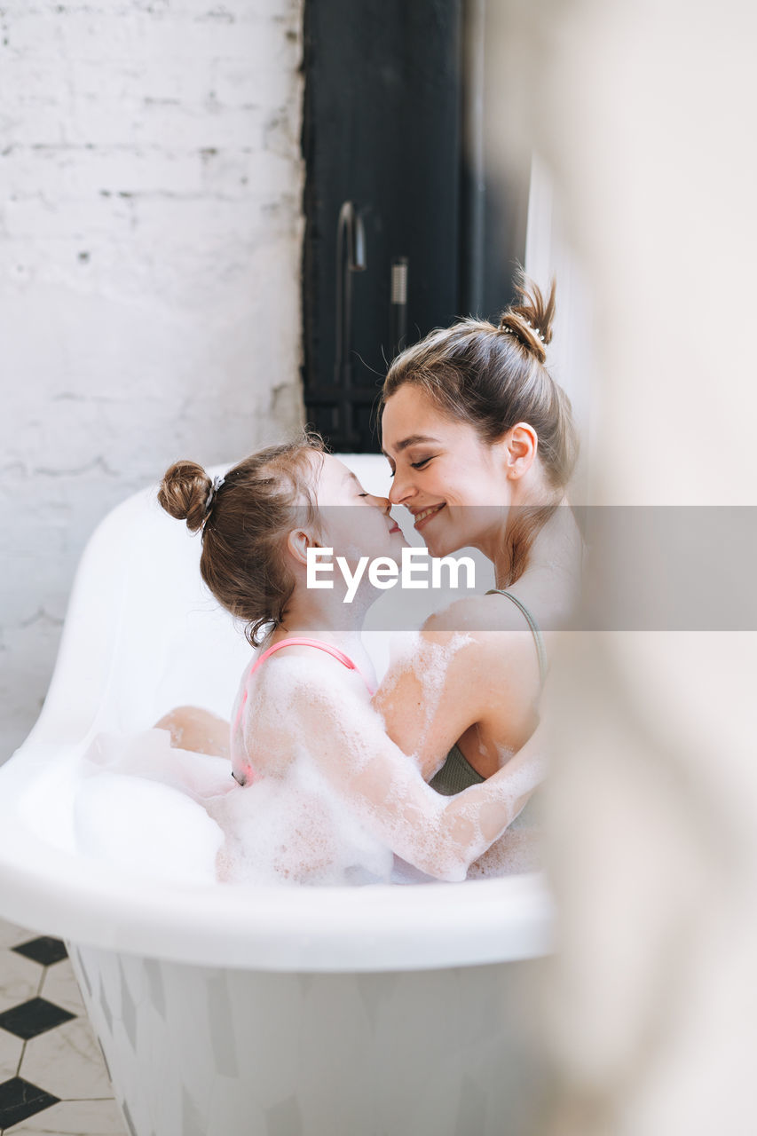 Young mother woman and little tween girl daughter having fun in bath with foam at home