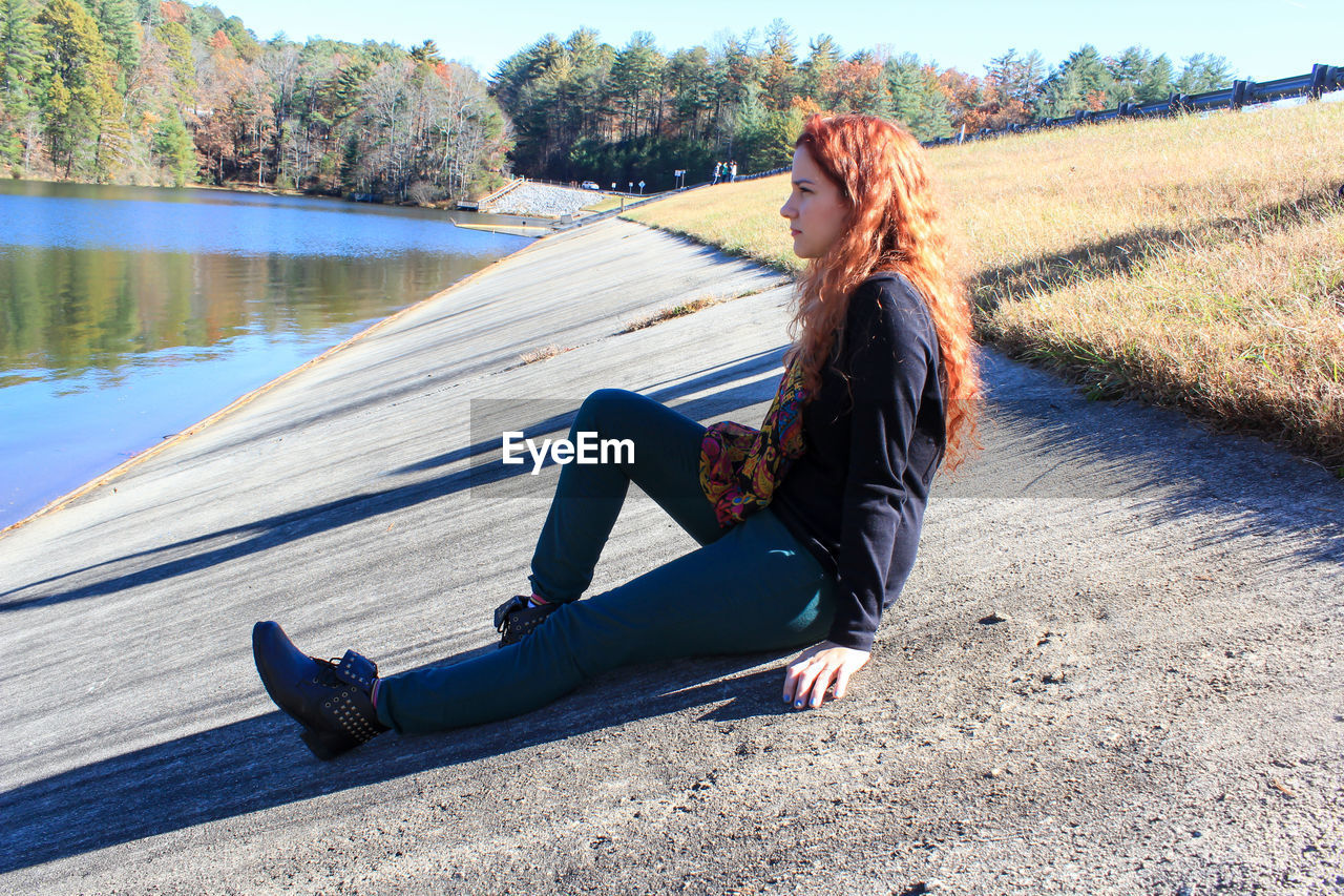 Young woman with red hair is sitting on the ground by a lake on a sunny but cold day georgia.