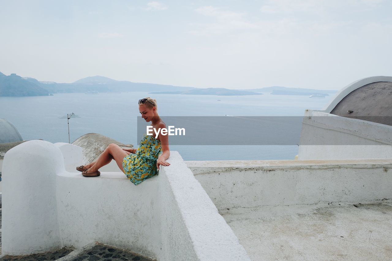 Young woman sitting on retaining wall by sea against sky