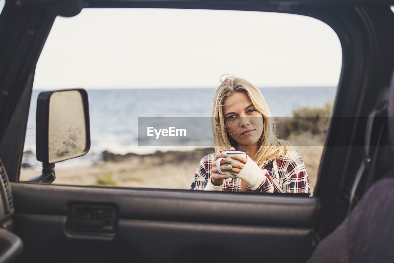 Portrait of young woman holding coffee cup by car against sky at beach