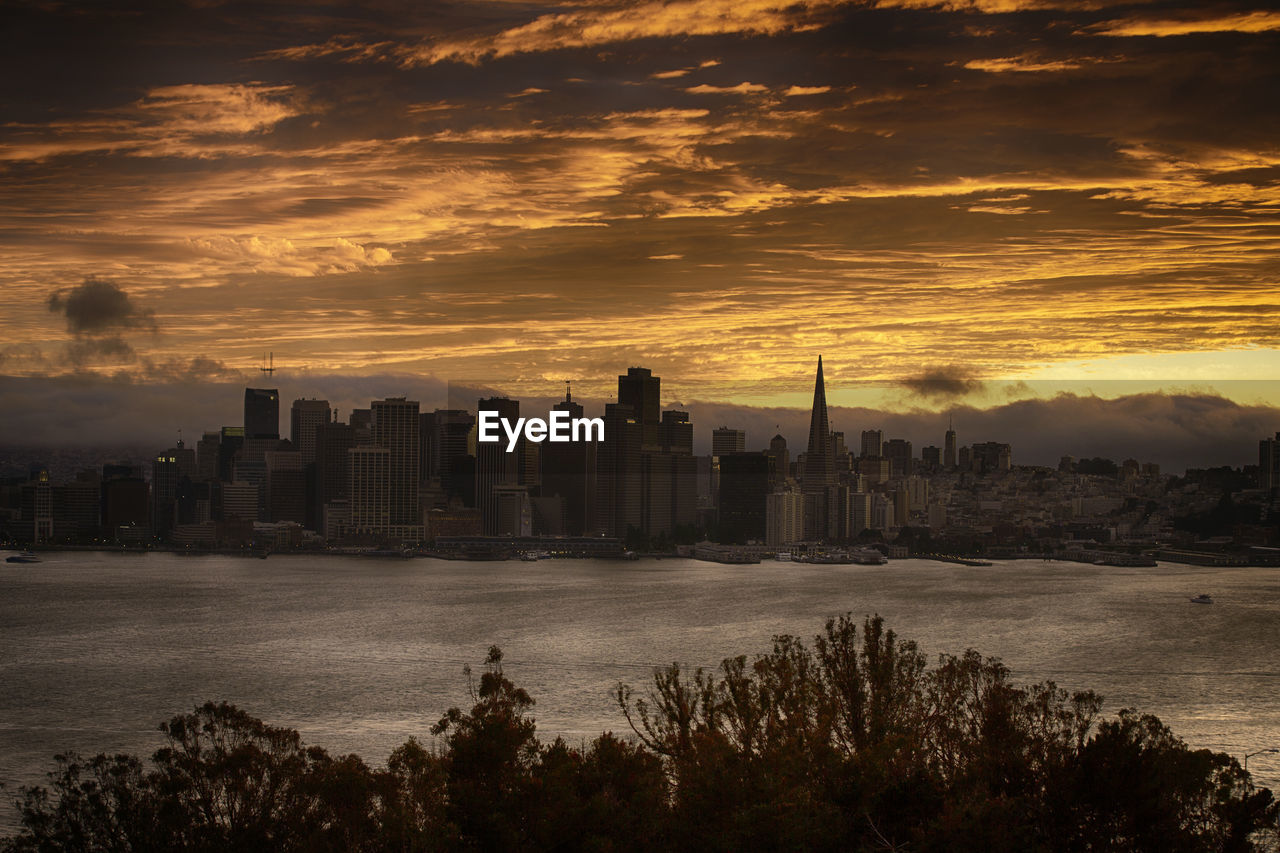 Urban skyline by sea against cloudy sky during sunset