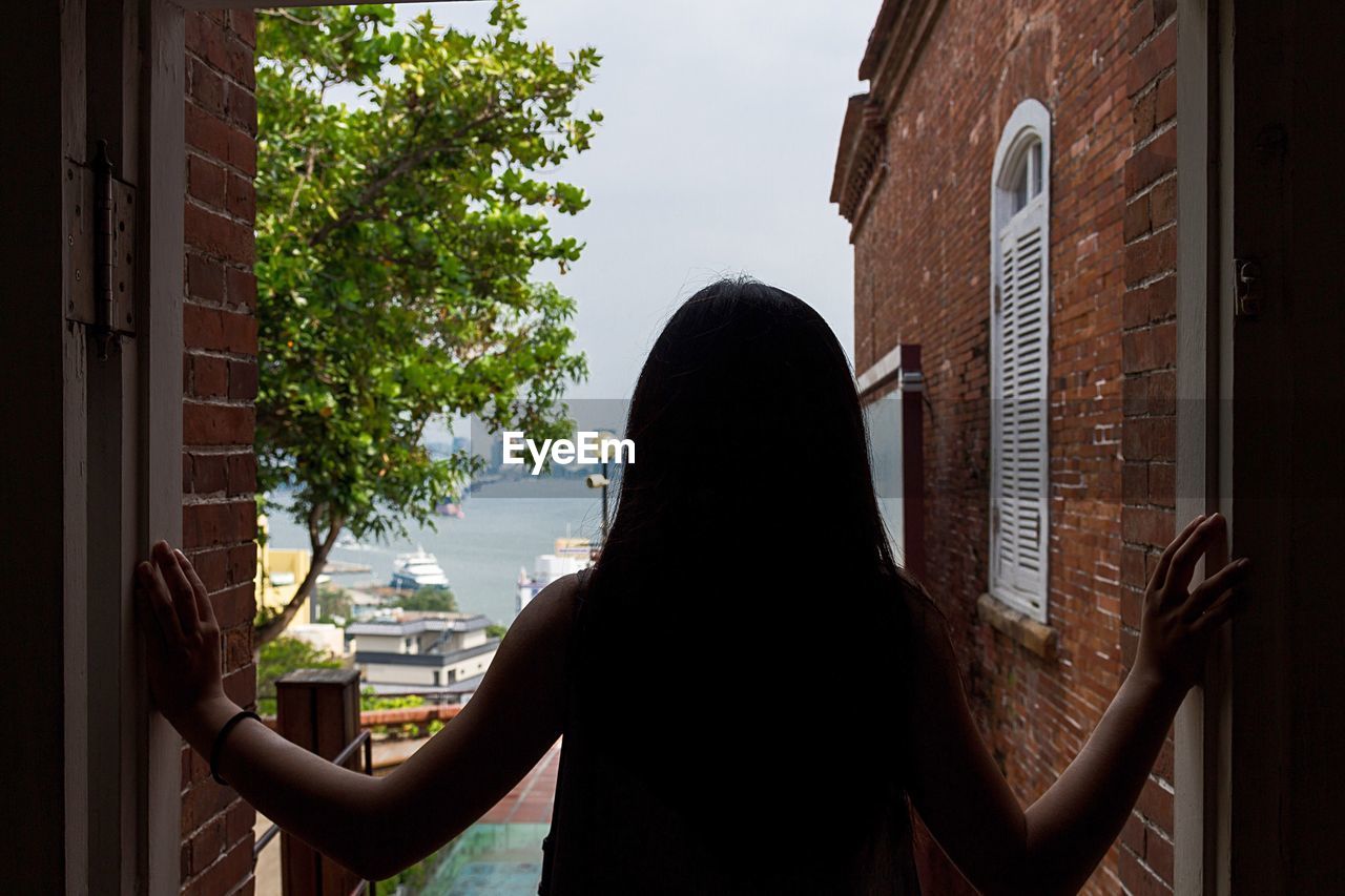 REAR VIEW OF WOMAN STANDING AGAINST WINDOW