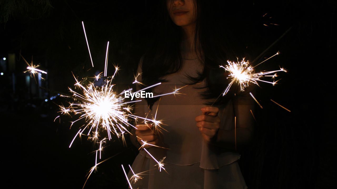 Midsection of teenage girl holding sparklers while standing outdoors at night