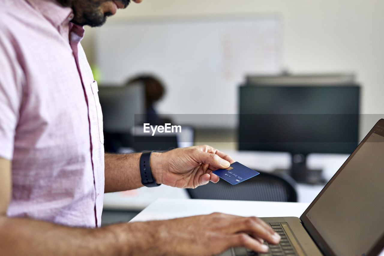 Midsection of businessman with credit card using laptop computer on desk in office