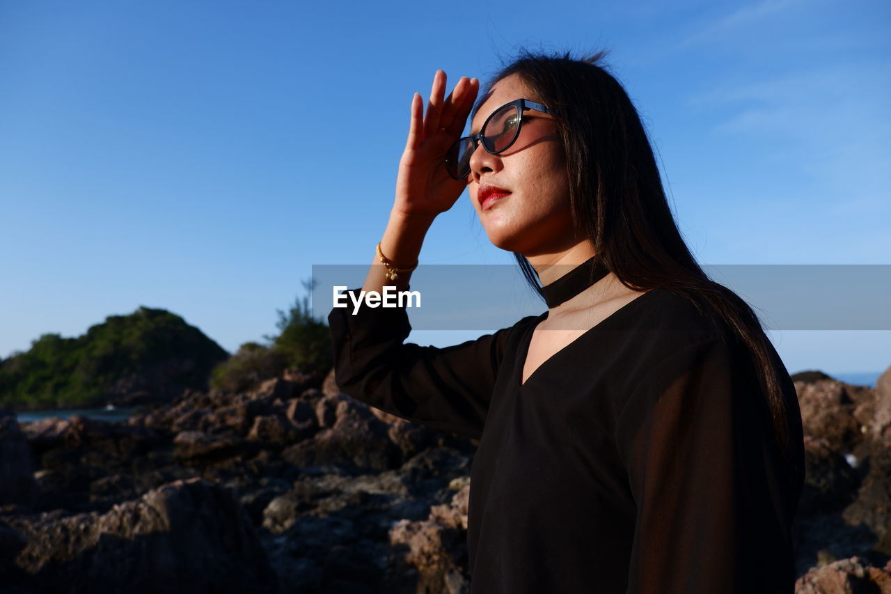Woman shielding eyes while standing by rock against sky