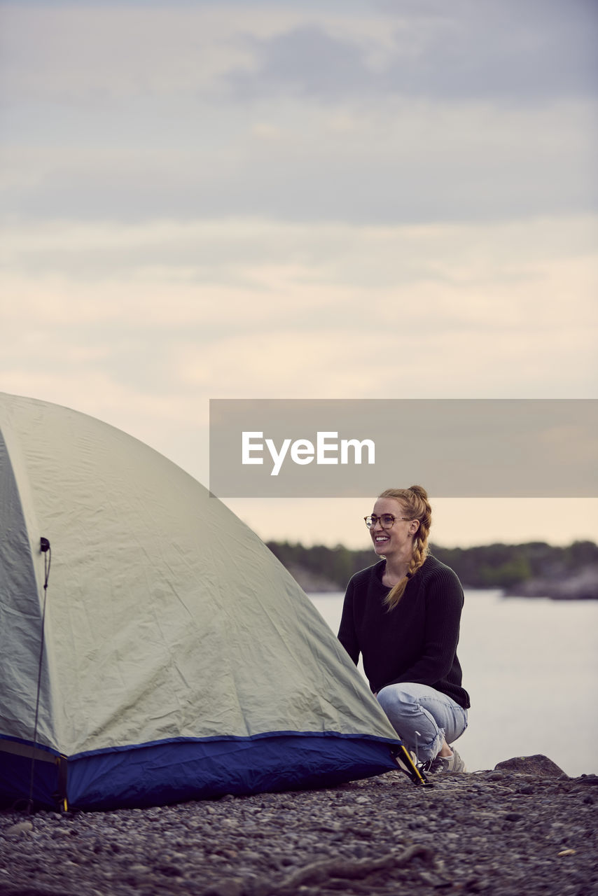 Smiling young woman crouching tent at beach against sky