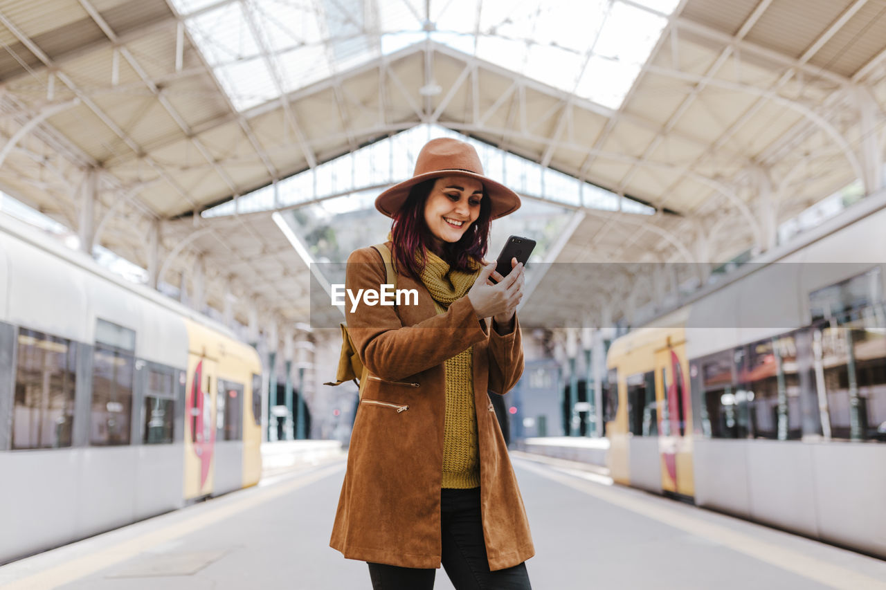 Smiling woman using smart phone while standing at railroad station platform