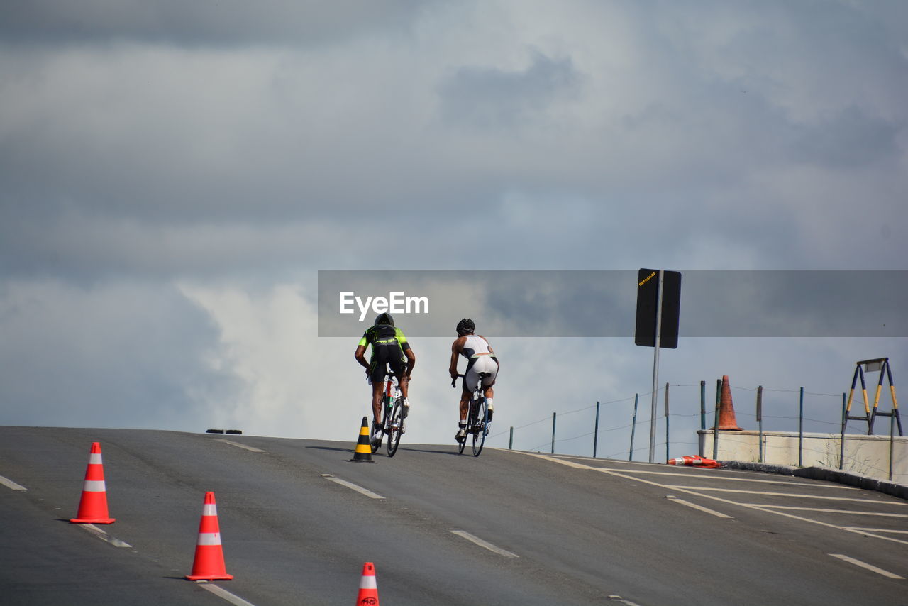 Rear view of friends riding bicycles on road against cloudy sky