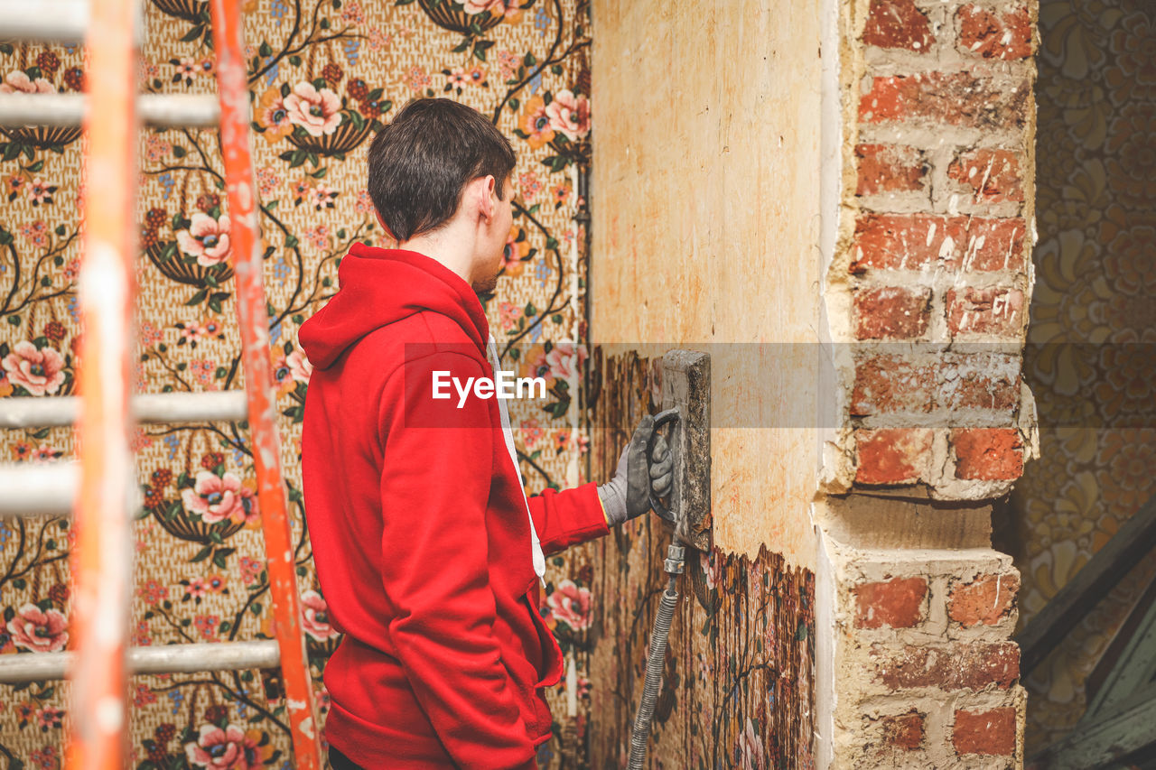 A young caucasian guy removes retro wallpaper using a steamer machine in an old abandoned house.