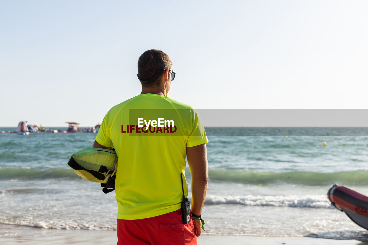Back view of anonymous male lifeguard in shorts and t shirt and maintaining safety on sandy seashore