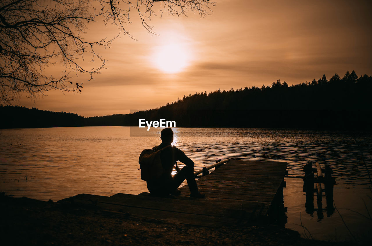 Silhouette man sitting by lake against sky during sunset