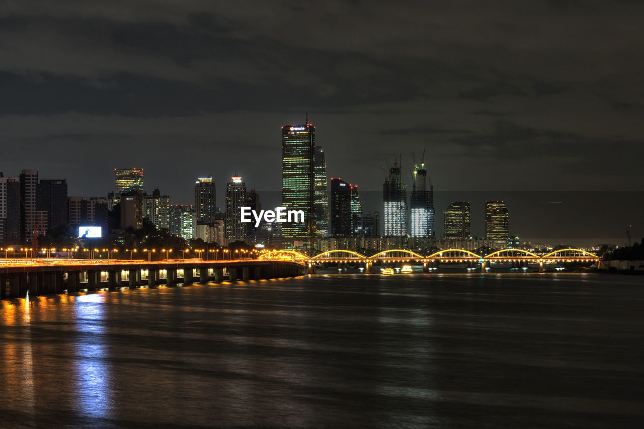 Illuminated buildings by river against sky at night