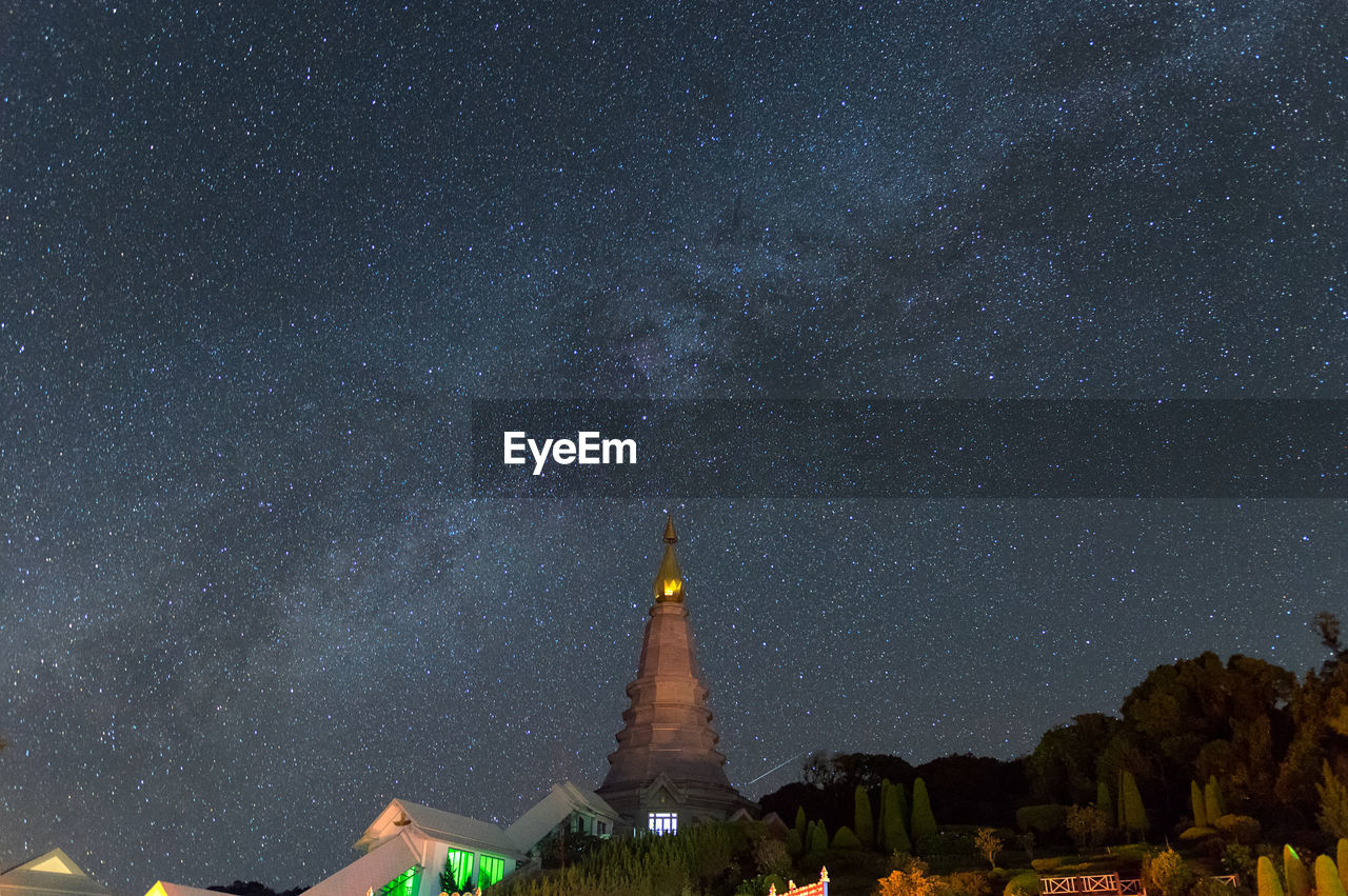 Low angle view of cross amidst buildings against sky at night milky way galaxy