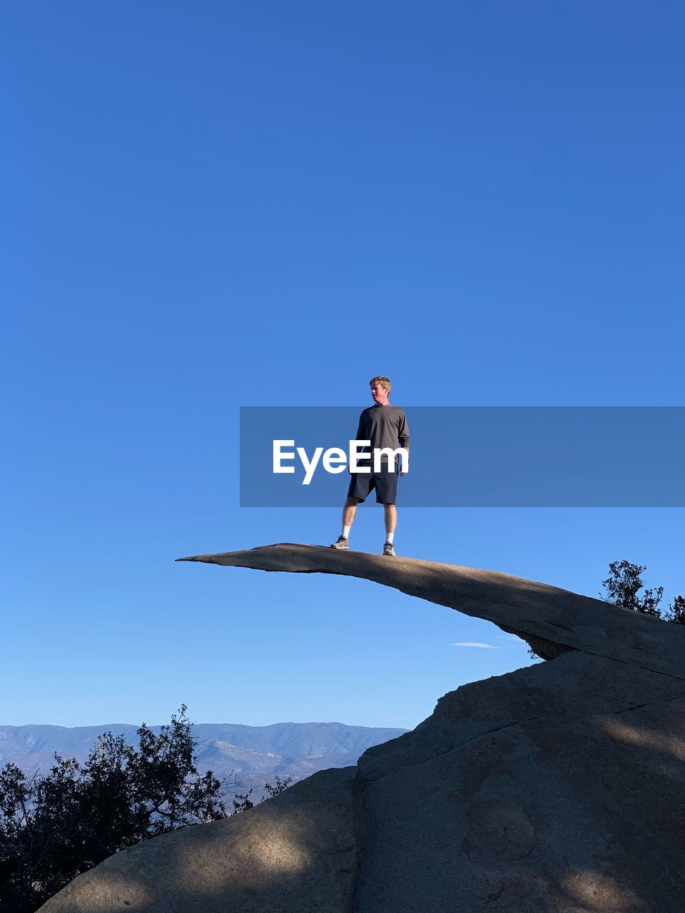 Low angle view of man standing on mountain ledge against clear blue sky