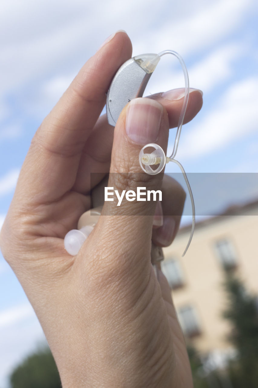 Close-up of hand holding hearing aid outdoors against sky