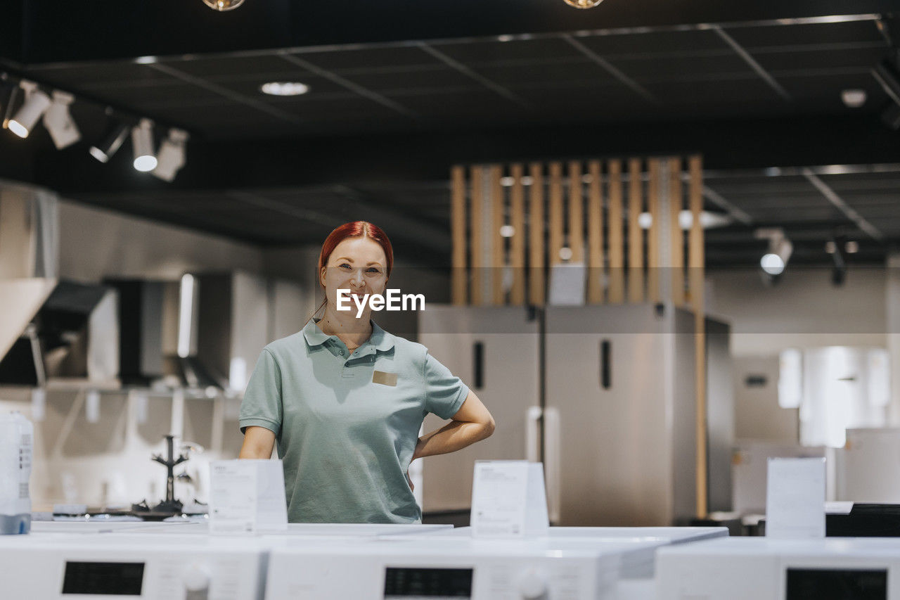 Portrait of smiling female retail clerk standing with hand on hip in electronics store