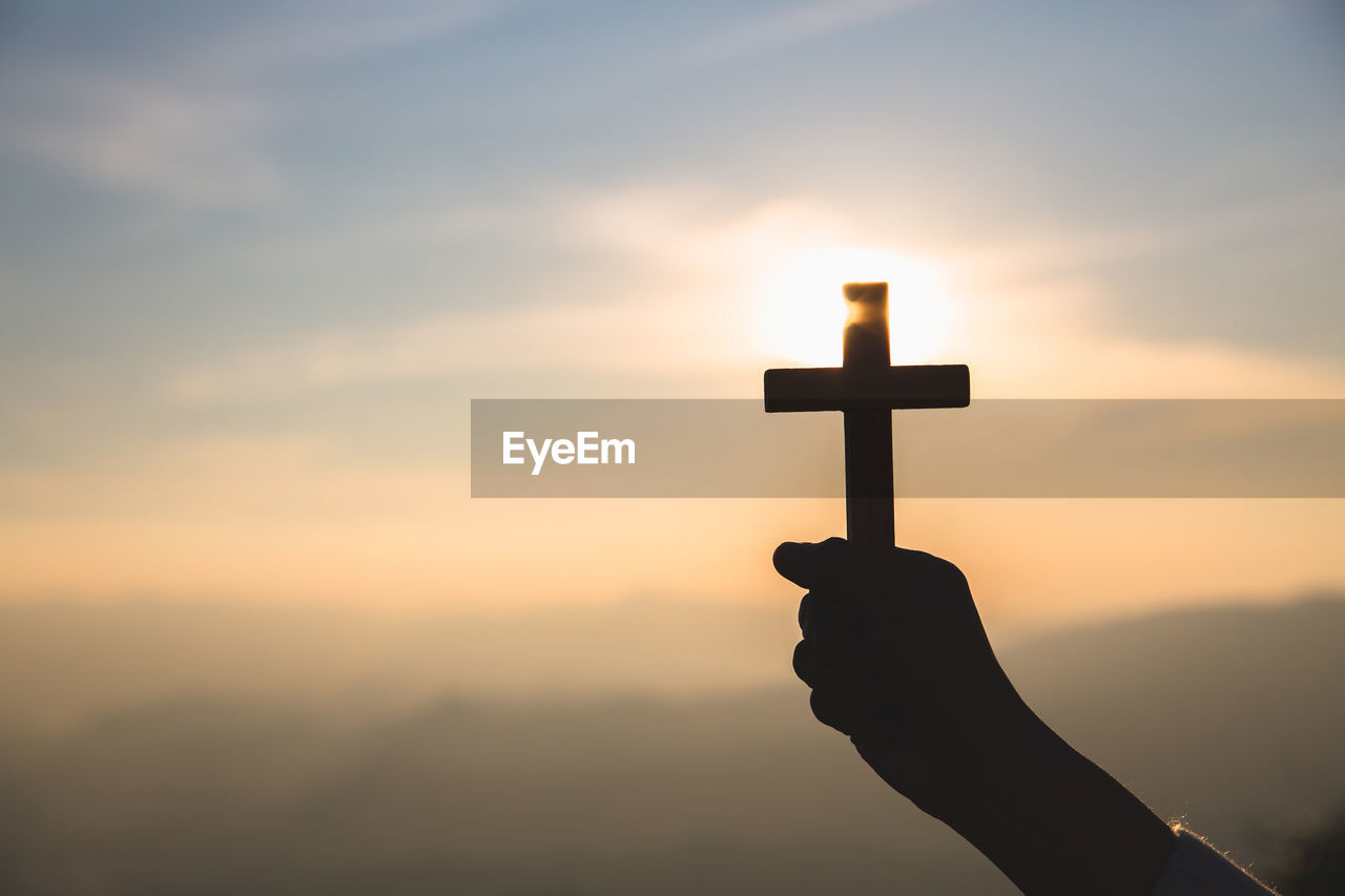 SILHOUETTE HAND HOLDING CROSS AGAINST ORANGE SKY