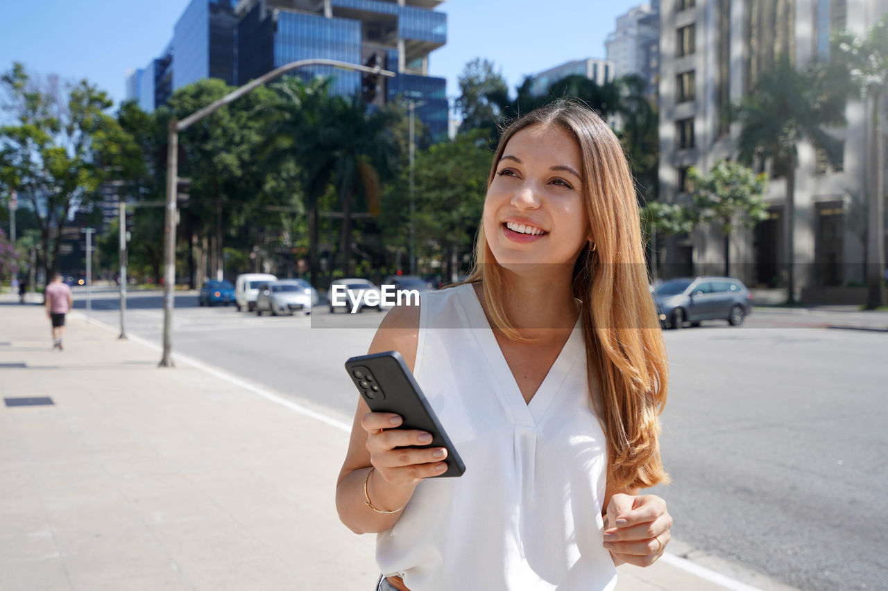 Beautiful young woman holding smartphone walking in financial district. 
