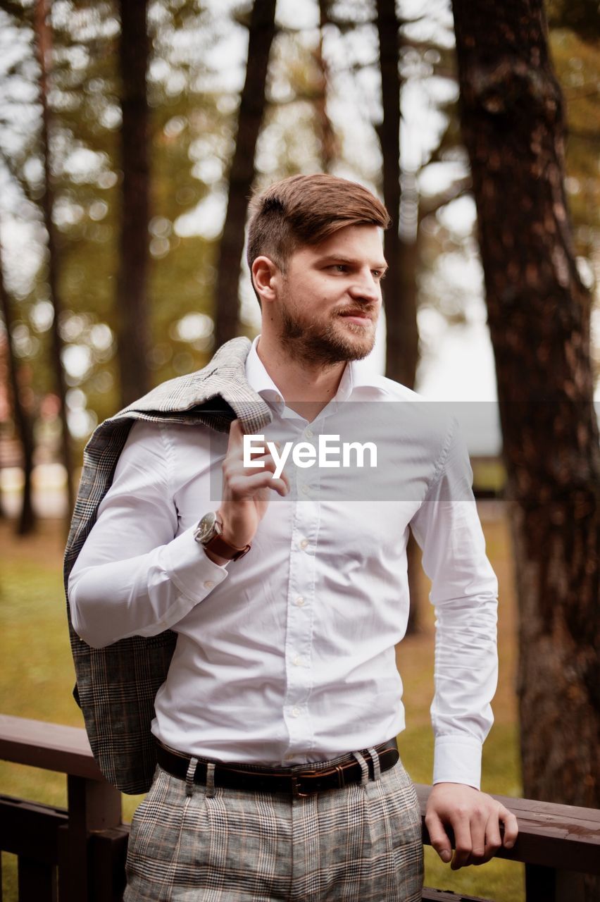 Young man looking away while standing by railing against trees in forest