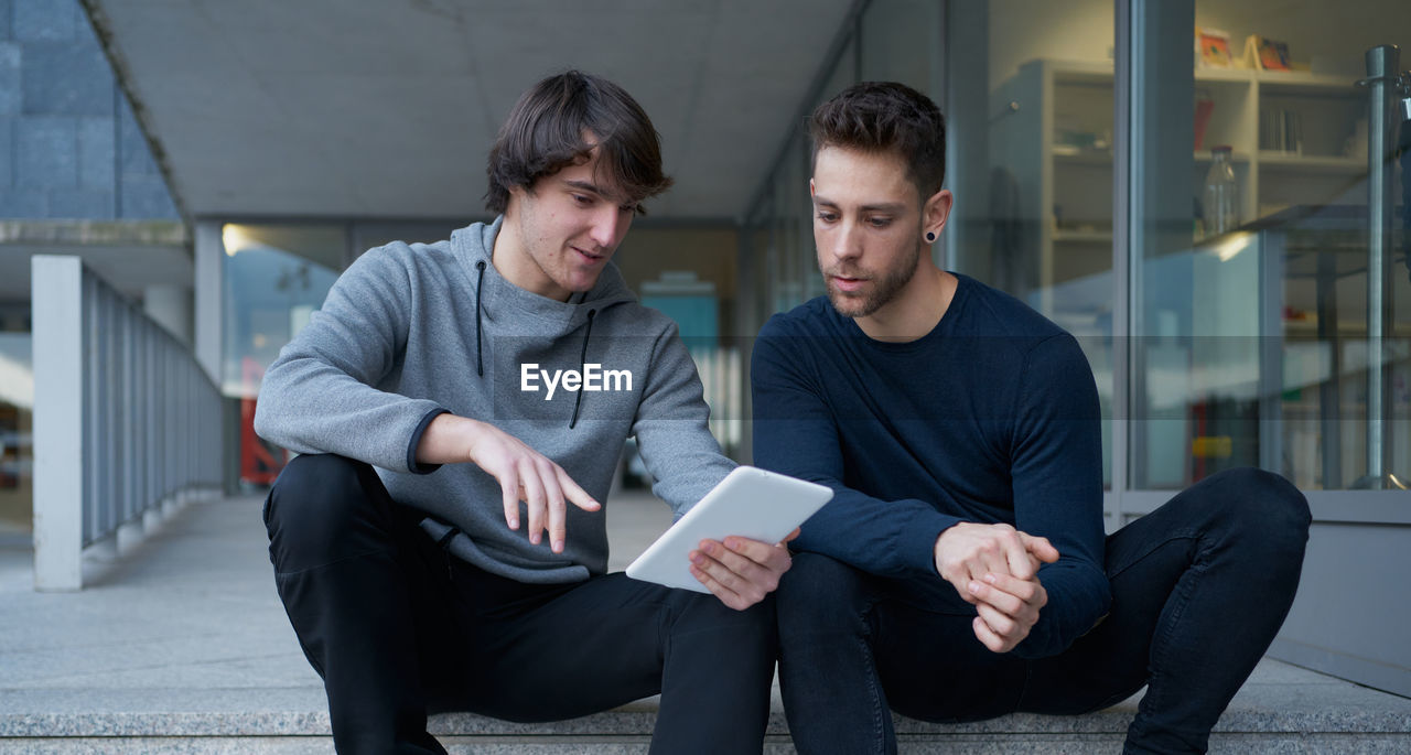 Front view of two young men talking sitting on a city staircase and lo