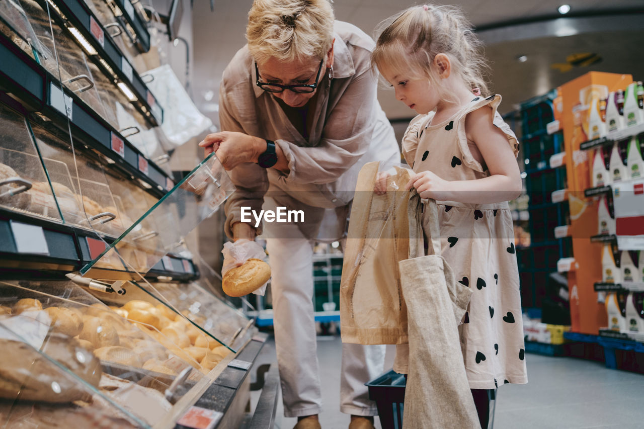 Senior woman buying bread with granddaughter holding bags at grocery store