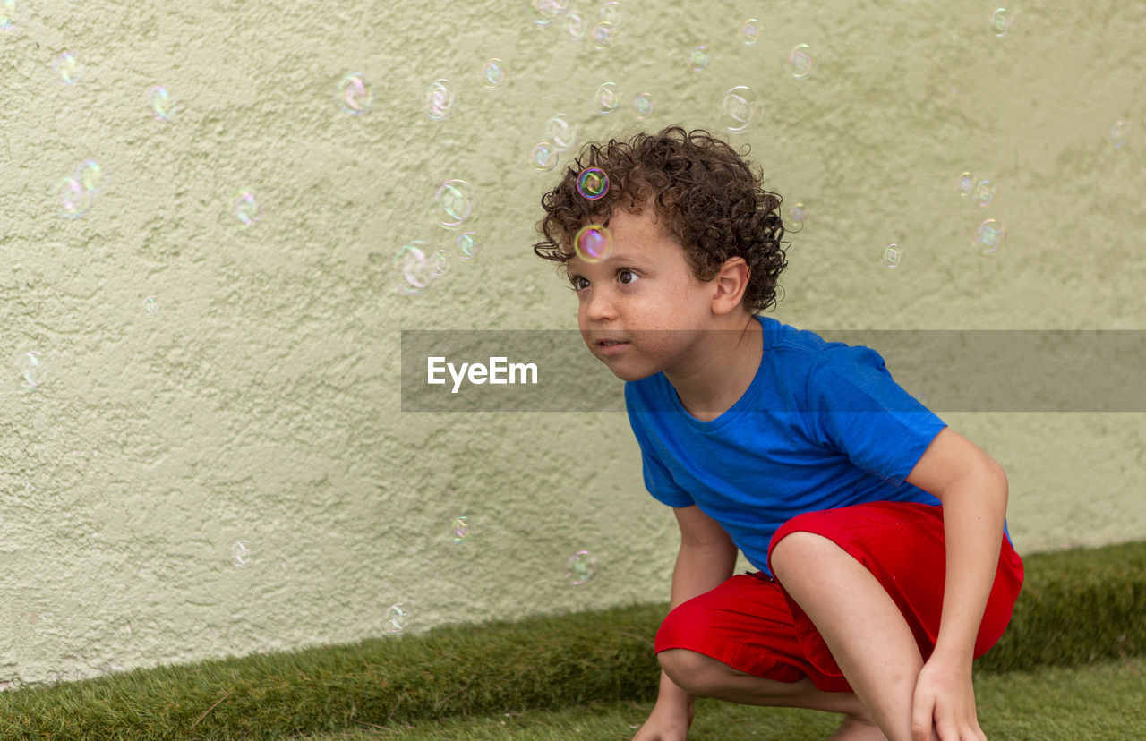 Curly-haired boy with freckles playing in the backyard with soap bubbles.