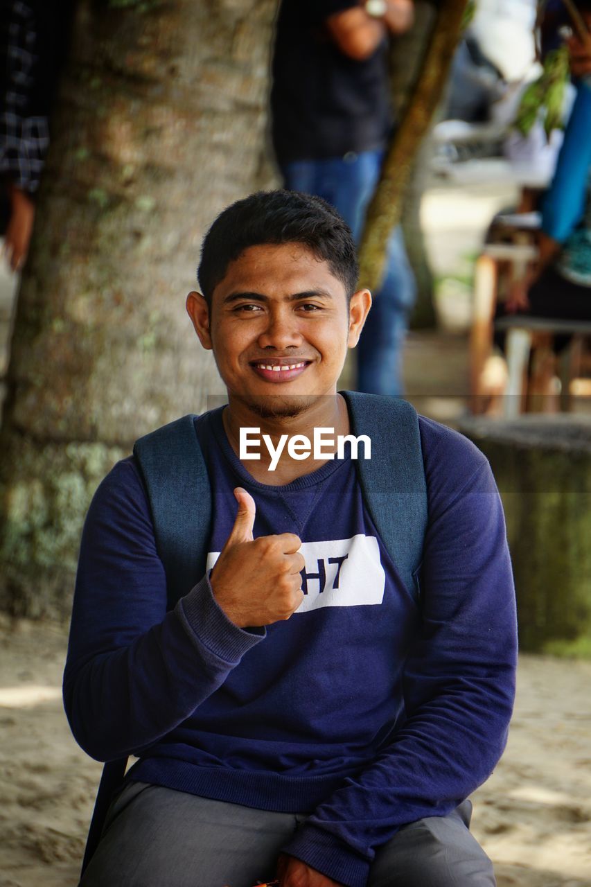 Portrait of smiling young man showing thumbs up against tree