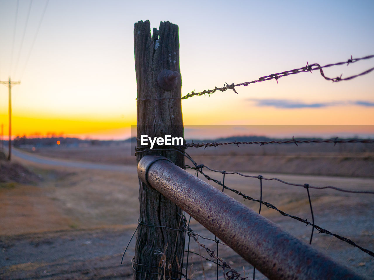 CLOSE-UP OF BARBED WIRE ON FIELD AGAINST SKY DURING SUNSET