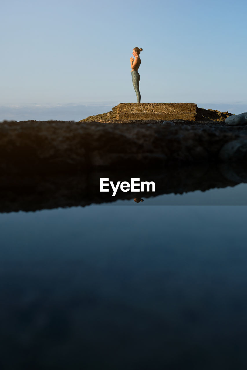 Full body side view of distant female in sportswear standing in mountain pose with prayer hands and meditating during yoga practice on rocky pier at seaside