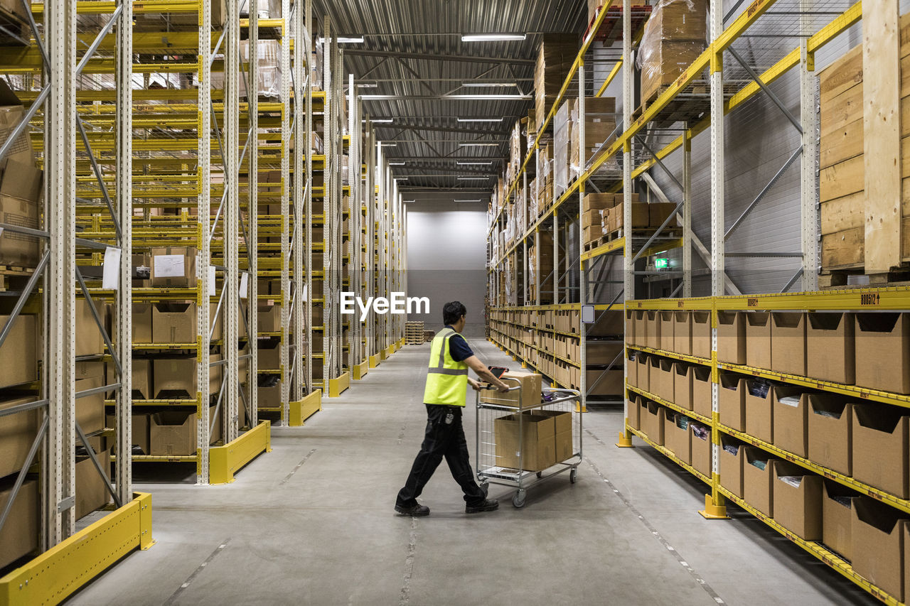 Full length of young warehouse worker pushing cart on aisle in industrial building