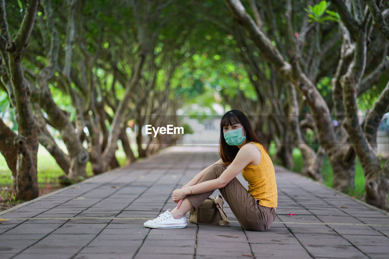 Portrait of young woman wearing mask and sitting by tree