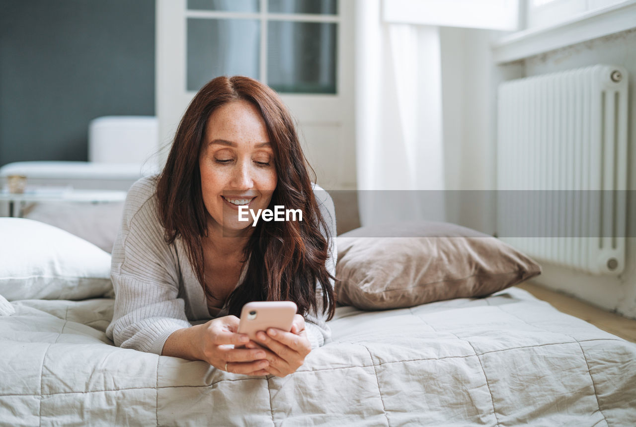 portrait of young woman using mobile phone while sitting on bed at home