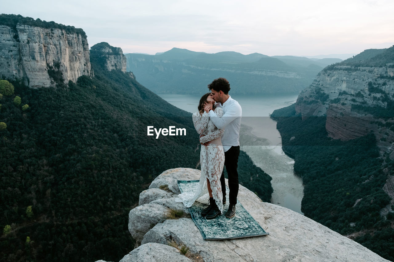 Side view of newlywed young man and woman in elegant clothes standing on top of rocky cliff of morro de labella embracing each other and kissing gently