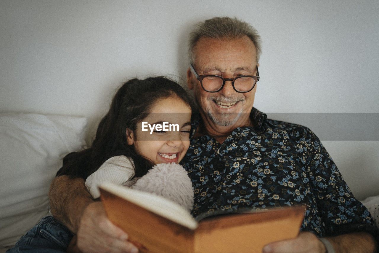Smiling senior man reading storybook with granddaughter on bed at home