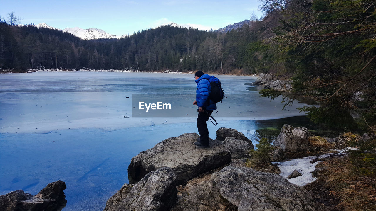 Side view of man standing on rock against frozen lake in forest